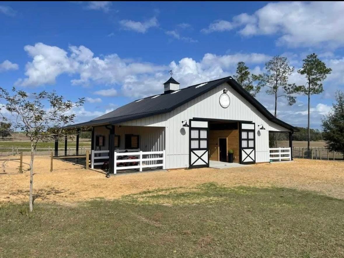 A white barn with a black metal roof and cupola on a sunny day, with exterior lights above the doors and a white fence.