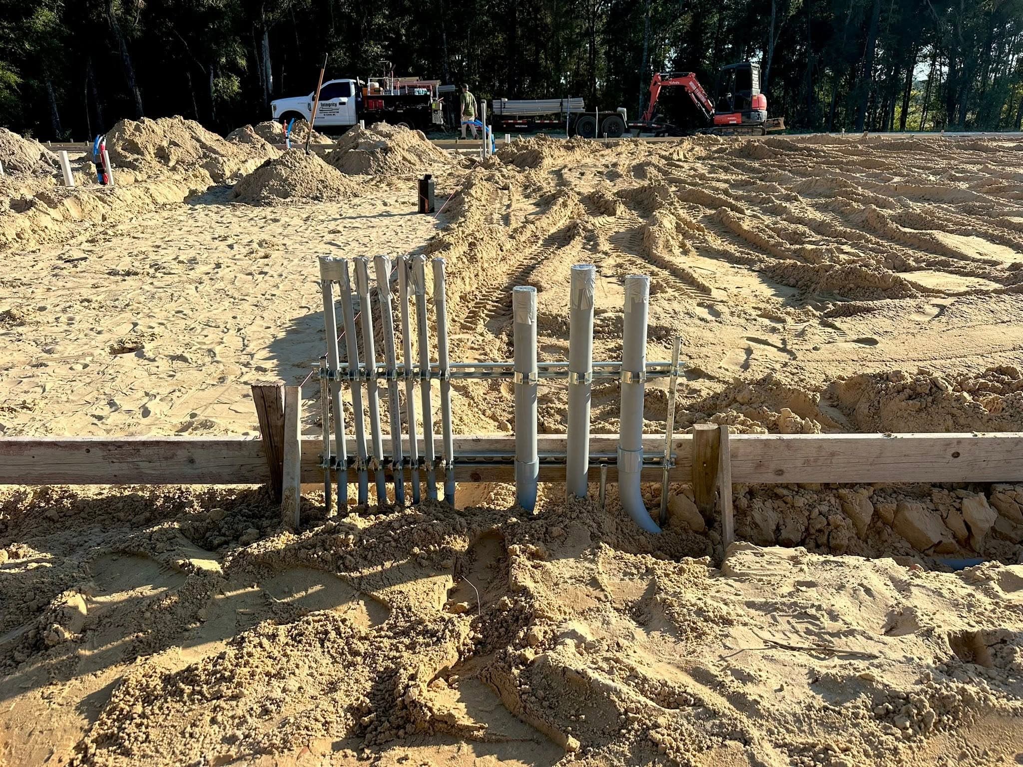 Rows of conduit stubs standing up out of the sand on a new construction site, with a truck, trailer, and excavator in the background.