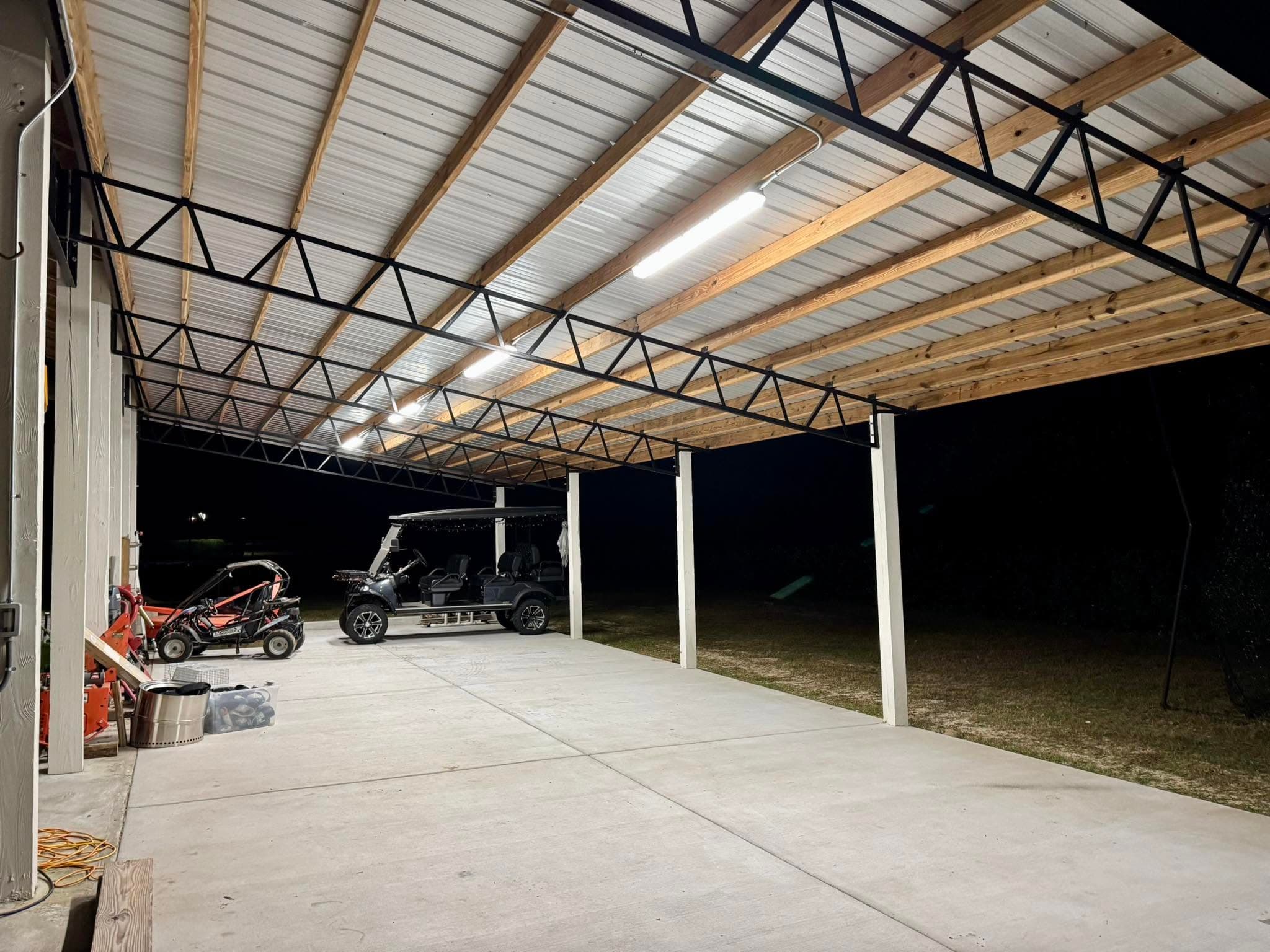 An open-sided covered barn at night with new strip lights on under a white metal roof and wood trusses, golf carts parked inside.