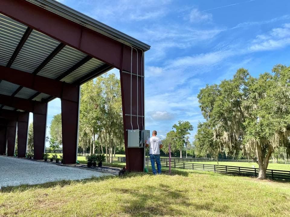 An Integrity crew member checking an electrical panel mounted on a red steel equestrian barn, with fenced pasture and live oaks behind him.