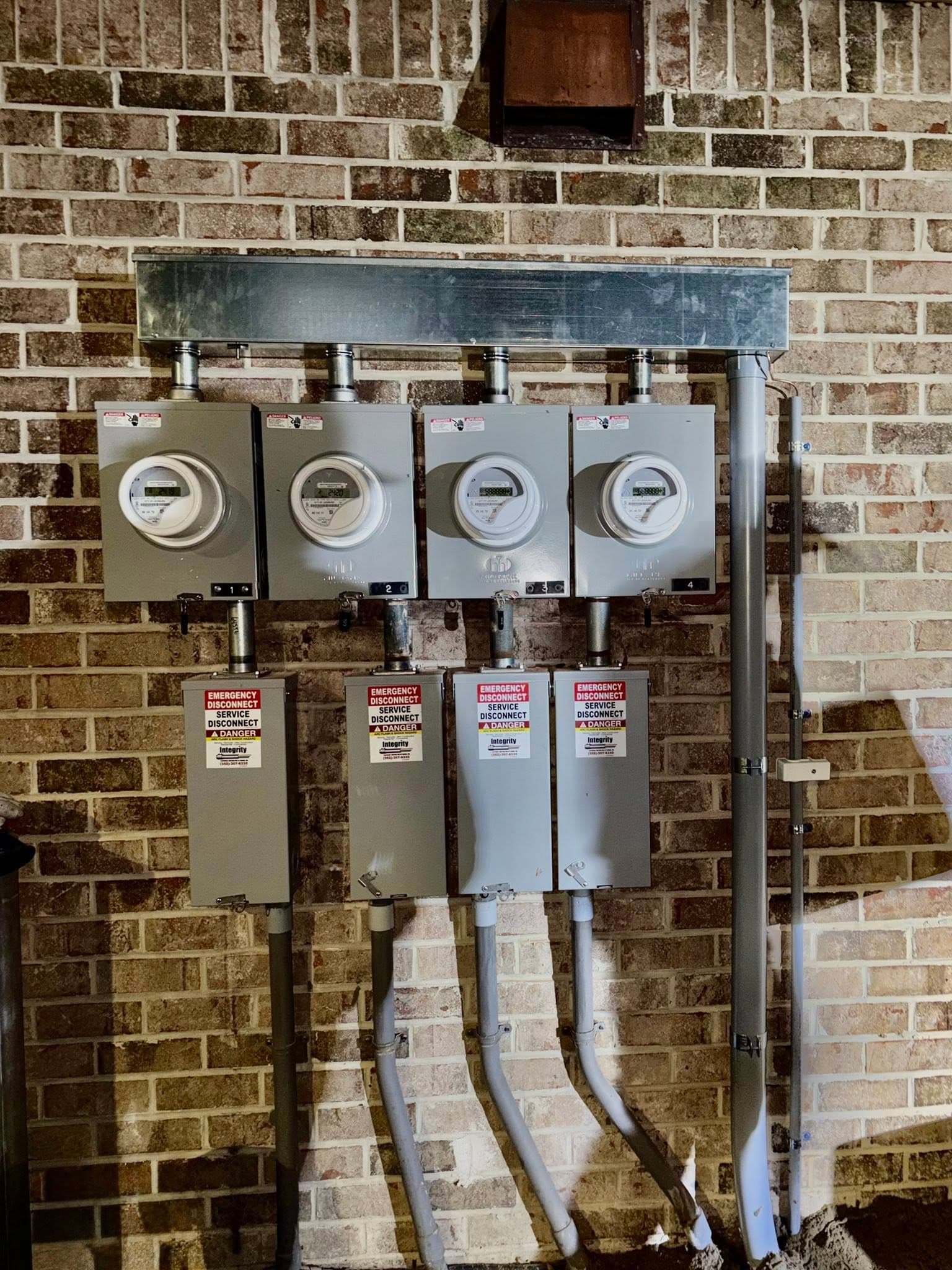 Four electric meters and four disconnect panels mounted in a row on a brick wall, conduit running down into the ground, photographed at night.