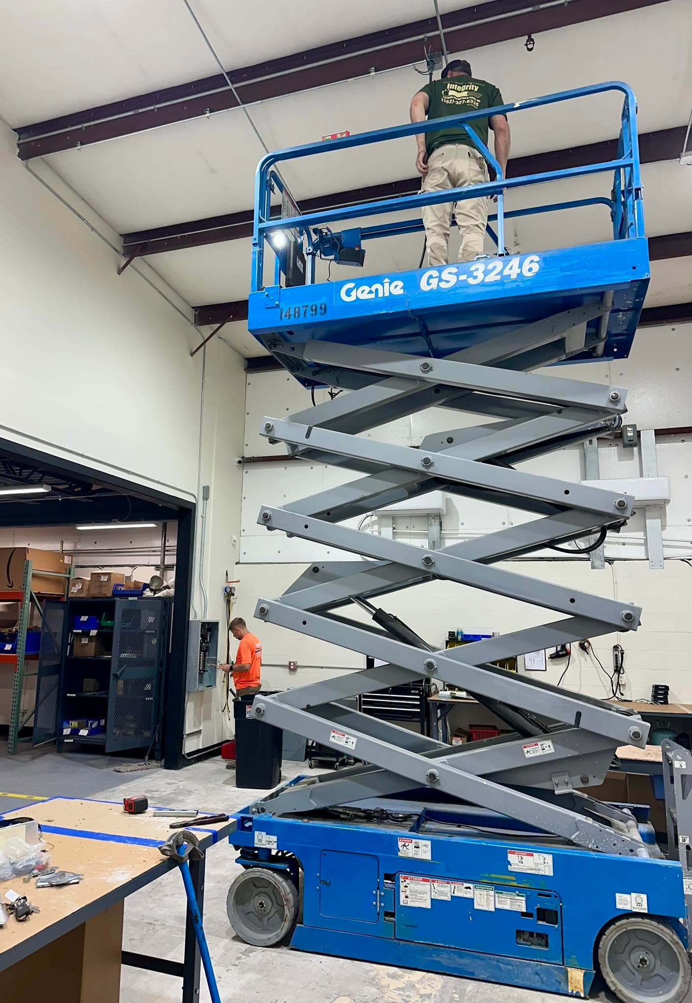A worker on a raised blue Genie scissor lift working near the ceiling inside a white metal building.