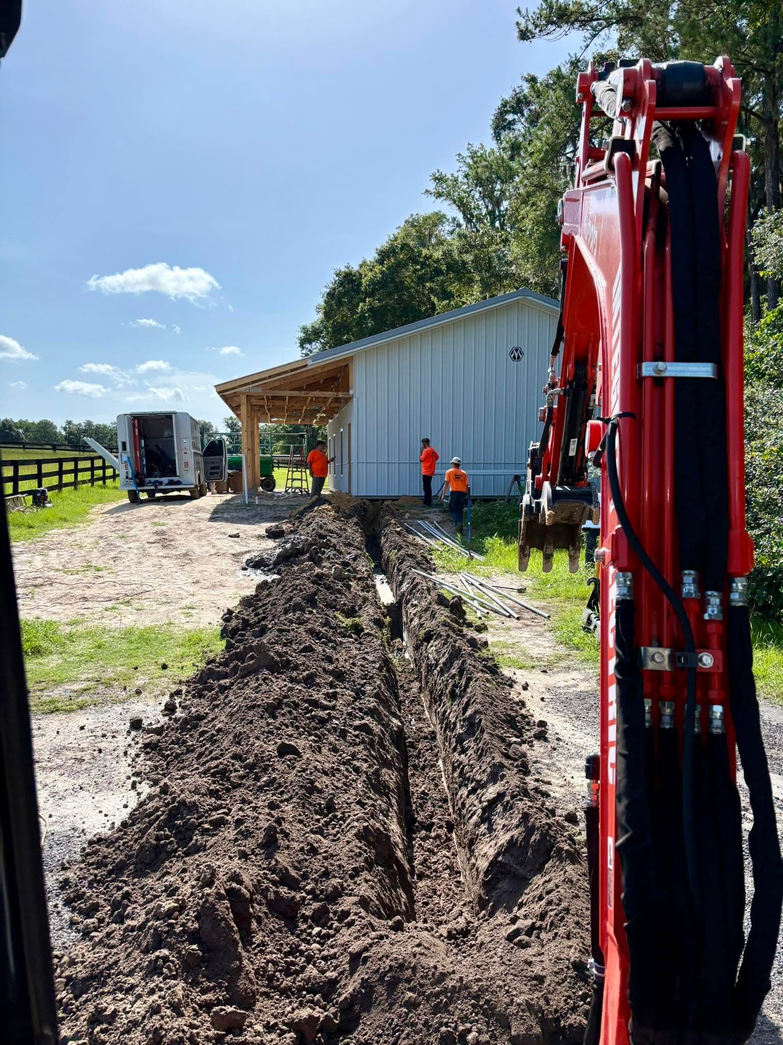 A red excavator digging a long trench across a grass field toward a white metal building, with a crew in orange shirts and a work truck in the background.