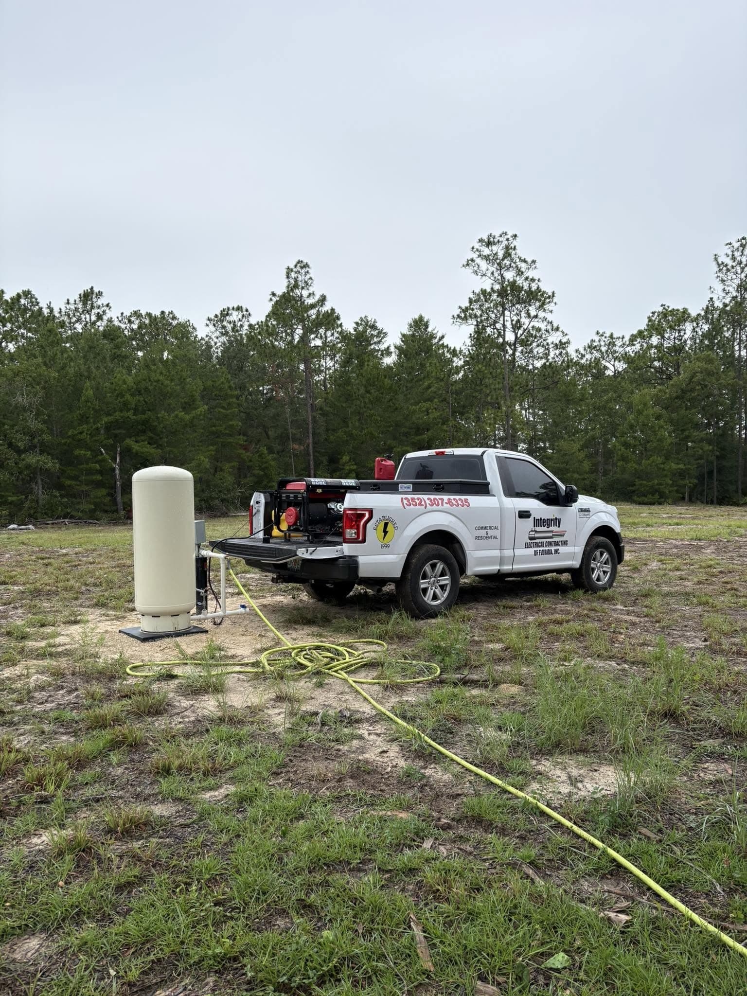 A white Integrity Electrical pickup truck parked at a rural job site next to a pressure tank, with yellow cable on the ground.