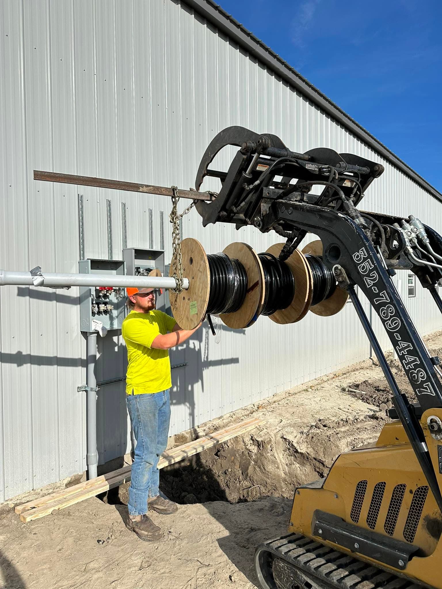 A worker in a yellow shirt and hard hat pulling wire from a large spool held up by a skid steer beside a white metal building.